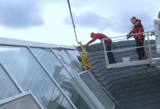 Zwei Arbeiter in Schutzkleidung der Glaserei Wuppertal stehen auf einer erhöhten Plattform und montieren bei bewölktem Himmel mit einer mechanischen Hebevorrichtung große Glasscheiben auf dem Schrägdach eines Gebäudes.