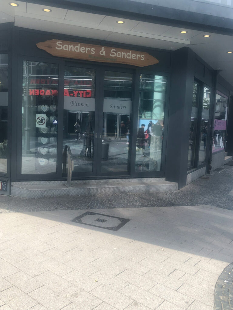 Ein Schaufenster in Düsseldorf mit einem Holzschild mit der Aufschrift "Sanders & Sanders". Auf den Schaufensteranlagen stehen die Worte "Blumen" und "Sanders", während Spiegelungen und Passanten das Glas über einem mit hellen und dunklen Steinen gepflasterten Bürgersteig beleben.