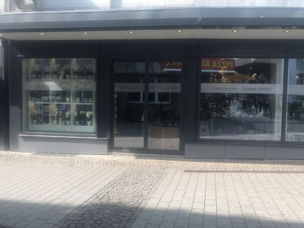 Ein Schaufenster in Düsseldorf mit großen gläsernen Schaufensteranlagen stellt Blumenarrangements aus. Auf den Schildern an den Fenstern steht Blumen Sanders und Eventfloristik. Die Straße davor ist mit grauen Pflastersteinen und einer Kopfsteinpflastereinfassung versehen.