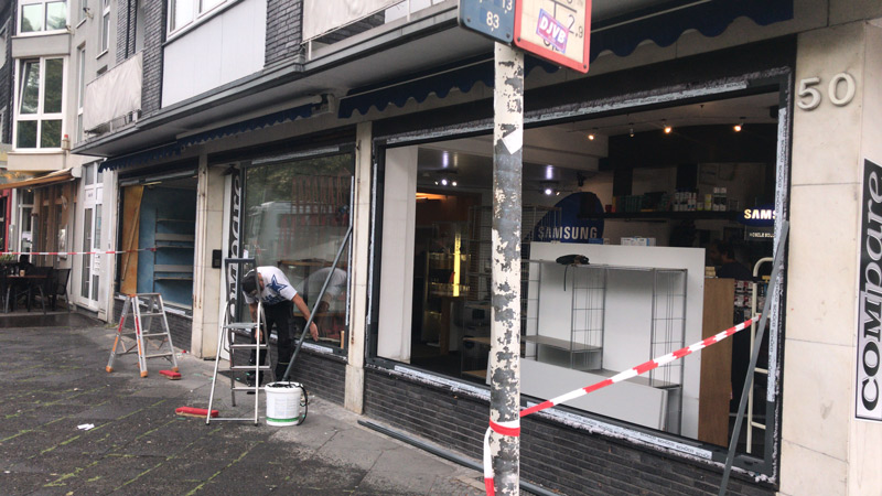 Eine Person bemalt den unteren Rahmen einer großen Schaufensteranlage in einer Stadtstraße. Rotes und weißes Klebeband sperrt den Bereich ab. In der Nähe stehen Leitern und ein Eimer. Im Inneren des Geschäfts sind Samsung-Produkte zu sehen.
