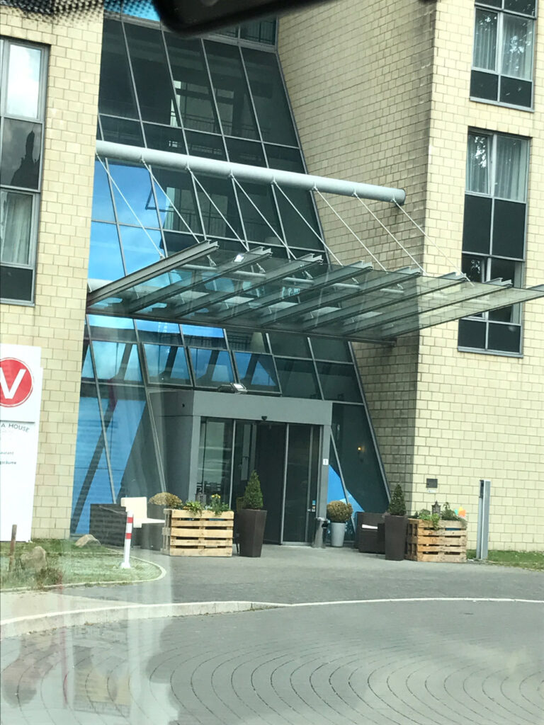 Das Bild zeigt den Eingang eines modernen Gebäudes mit einem großen Glasvordach und beigen Ziegelwänden, auf dem Schaufensteranlagen Wuppertal abgebildet sind. Topfpflanzen flankieren die Türen, während davor ein Kreisverkehr und ein Schild zu sehen sind.