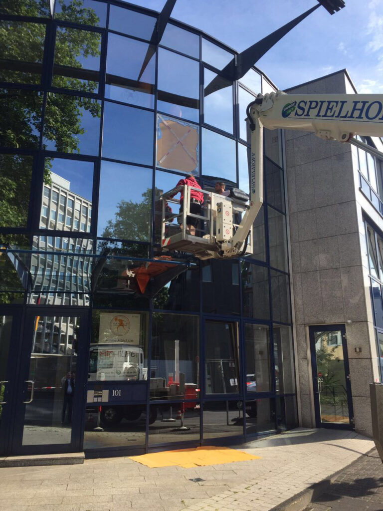 Zwei Arbeiter auf einer erhöhten Plattform reparieren oder montieren Glasscheiben an einem modernen Gebäude, das Teil der Schaufensteranlagen Wuppertal ist. Ein weißes Fahrzeug mit der Aufschrift SPIELHOFF auf dem Kranarm stützt die Plattform, während sich Bäume und Gebäude im Glas spiegeln.