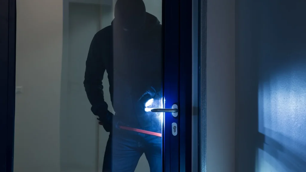 Eine dunkel gekleidete Person mit Kapuze bricht nachts mit einer Taschenlampe und einem Brecheisen durch eine Glastür in ein Gebäude ein und unterstreicht damit die Bedeutung des Einbruchschutzglases Velbers-Neviges für mehr Sicherheit.