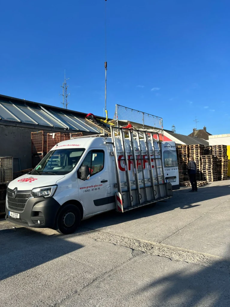 Ein weißer Kleintransporter mit GRAEFE-Schriftzug und Glasträgern steht vor einem Industriegebäude in Wuppertal neben Palettenstapeln. Eine Person steht in der Nähe, und Glasfensterscheiben glitzern unter dem klaren blauen Himmel. Gräfe Glas und Fenstertechnik in Wuppertal