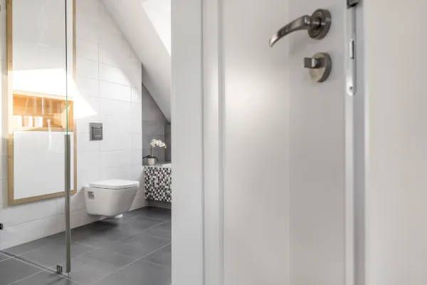 Das moderne Badezimmer in Velbert-Neviges ist mit grauen Bodenfliesen, einer Wandtoilette, einer Duschenabtrennung aus Glas und einem gemusterten Waschtisch mit weißem Waschbecken ausgestattet; der Blick wird teilweise durch eine offene weiße Tür mit einem Metallgriff verdeckt.