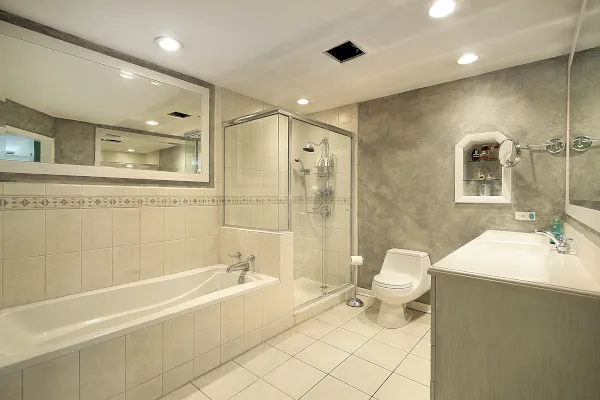 Modernes Bad in Velbert-Neviges mit großer Badewanne, gläsernen Duschen, Toilette und hellem Waschtisch. Neutrale Fliesen bedecken den Boden und die Wände, über dem Waschbecken befindet sich ein großer Spiegel und die Deckenbeleuchtung ist eingelassen.