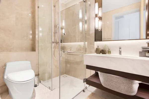 Modernes Badezimmer in Neviges, Velbert, mit beigen Marmorwänden, einer Duschenabtrennung aus Glas, einer weißen Toilette, einem großen Spiegel, moderner Beleuchtung, einem weißen Waschbecken mit Ablage darunter und dekorativen Gegenständen auf der Arbeitsplatte.