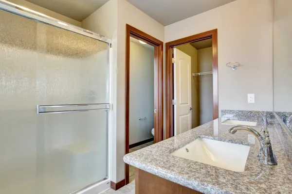 Modernes Badezimmer mit Glasdusche mit Schiebetüren, Waschbecken mit Granitplatte und Wasserhahn, großem Spiegel und Blick in einen kleinen Raum mit Toilette und begehbarem Kleiderschrank - perfekt für elegantes Wohnen in Neviges.