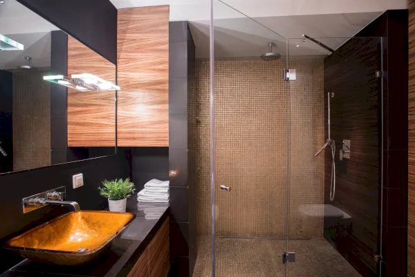 Ein modernes Bad mit Holzschränken, einem orangefarbenen Glaswaschbecken, einer Wandarmatur, einer kleinen Topfpflanze, gestapelten Handtüchern und einer begehbaren Glasdusche mit Glastüren und braunen Mosaikfliesen - perfekt für stilvolles Wohnen in Wuppertal.