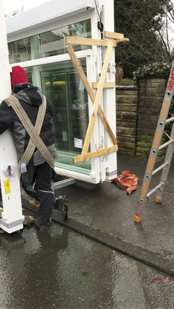 Eine Person in Outdoor-Kleidung sichert Glasfensterscheiben in der Nähe einer Leiter auf nassem Pflaster neben einer Steinmauer, wahrscheinlich um neue Fenster für den Einbau in Düsseldorf vorzubereiten.