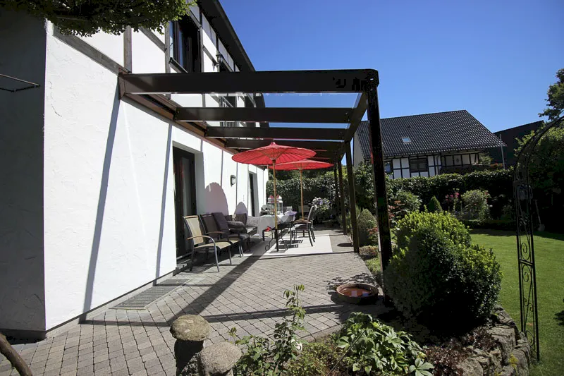 An ein weißes Haus in Hilden ist eine moderne Terrasse mit Glasfensterscheiben, einem Glasdach, einer Essgruppe im Freien und einem roten Sonnenschirm angebaut. Die Terrasse blickt auf einen gepflegten Garten mit üppigem Grün und Nachbarhäusern im Hintergrund.