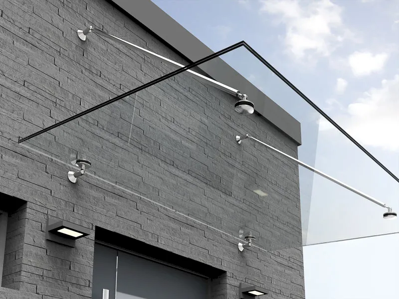 Eine moderne Gebäudefassade in Hilden zeigt ein rahmenloses Glasvordach aus Glasfensterscheiben, das von Metallstäben über dem Eingang getragen wird, vor einer grauen Backsteinwand und einem teilweise bewölkten Himmel.