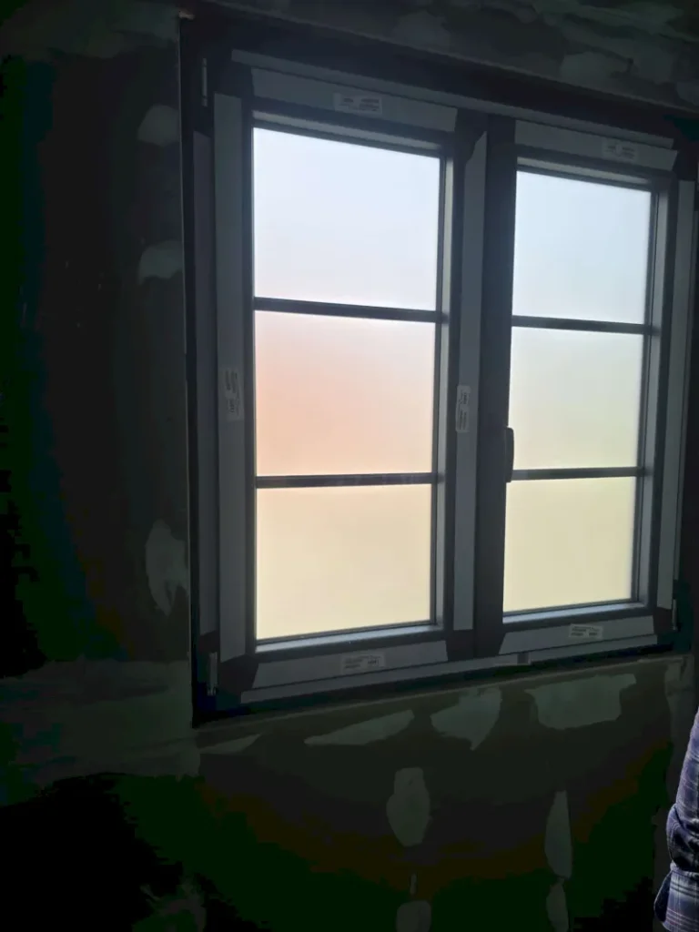 Eine geschlossene Glasfensterscheibe mit Milchglas lässt diffuses Licht herein, das in eine Wand mit sichtbaren Flecken unfertigen Putzes eingelassen ist. Am rechten Rand ist ein Teil der Schulter einer Person zu sehen, was ein an Wuppertal erinnerndes Ambiente hervorruft.