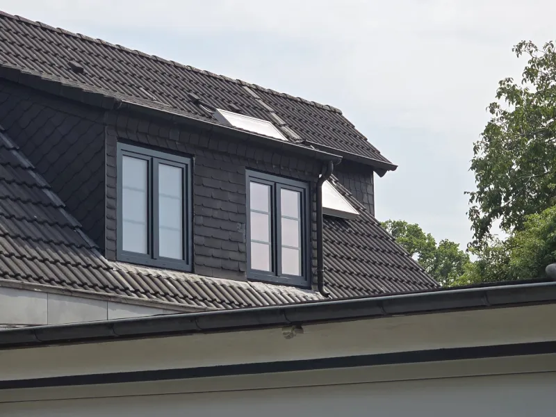Dunkelgraues Ziegeldach mit zwei Dachgauben mit Glasfensterscheiben; über einem Fenster befindet sich ein Solarpanel. Im Hintergrund sind grüne Bäume und der wolkenverhangene Himmel von Wuppertal zu sehen.