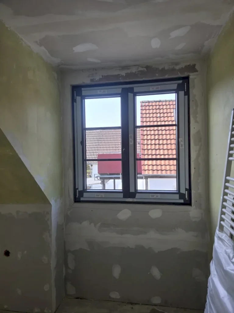 Ein zu renovierender Raum mit unfertigen Wänden und einer großen Glasfensterscheibe. Das Fenster ist geöffnet und gibt den Blick auf die Dächer der benachbarten Gebäude in Wuppertal frei. Auf der rechten Seite des Bildes befindet sich ein weißer Heizkörper.