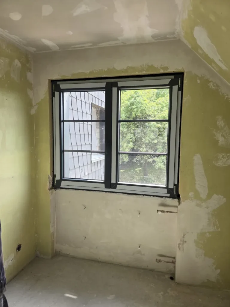 Ein halbfertiger Raum mit ungestrichenen, geflickten Wänden und Glasfensterscheiben im Fenster mit Blick auf grüne Bäume und ein angrenzendes Gebäude in Wuppertal. Der schwarze Fensterrahmen ist außen mit einem Schutznetz versehen.