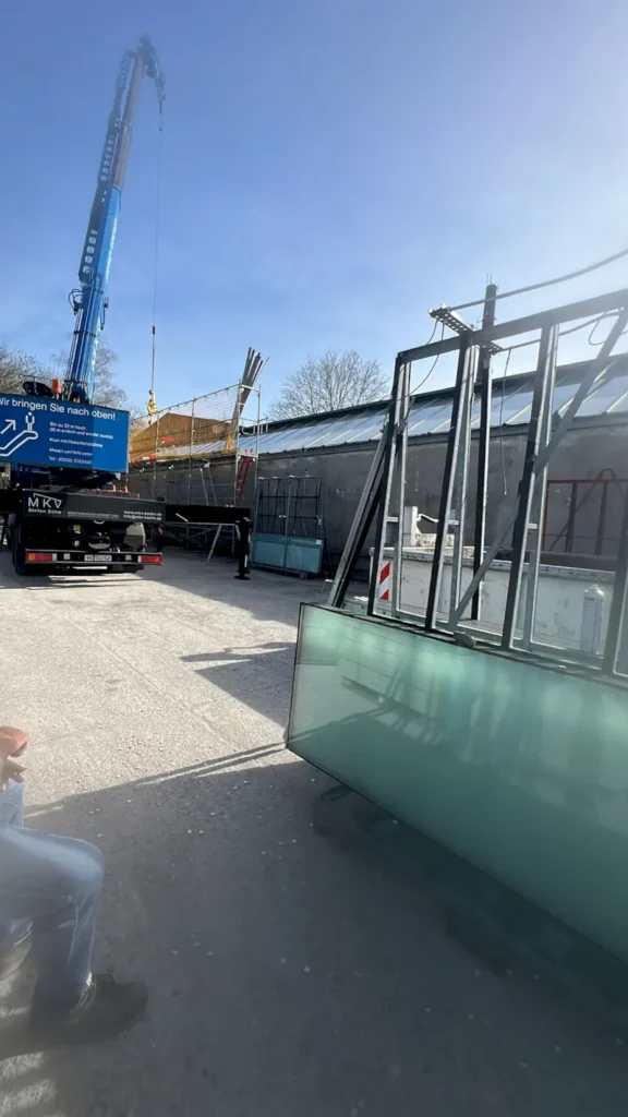 Eine Baustelle in Wuppertal mit einem Kran, der Material anhebt, Glasfensterscheiben, die auf Regalen gestapelt sind, und einem in der Nähe geparkten Lastwagen; eine Person sitzt in der linken unteren Ecke, Sonnenlicht wirft Schatten auf den Boden.