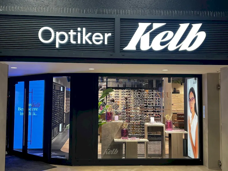 Schaufenster von "Optiker Kelb" in Wuppertal mit Leuchtreklame, großen Glasfensterscheiben mit Brillen und einem Plakat einer Brillenträgerin im Inneren. Der Eingang ist hell erleuchtet und einladend.