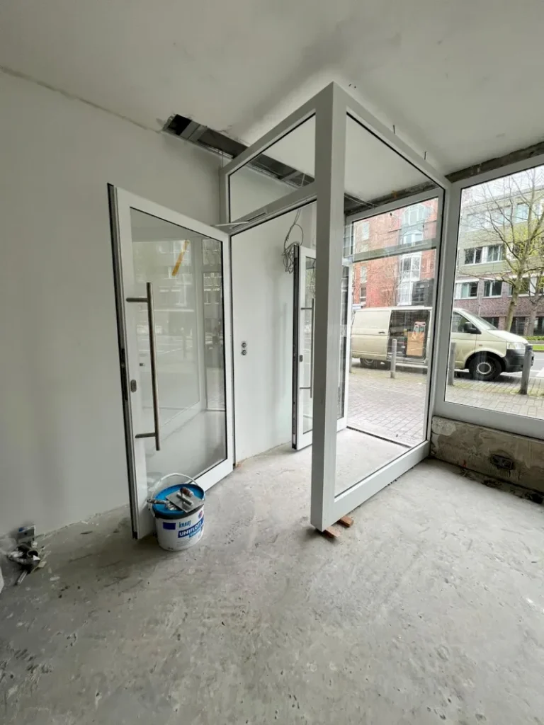 Eine Glastür und ein Rahmen für Glasfensterscheiben sind im Innenraum installiert, aber die seltsam gedrehten Rahmen scheinen nicht richtig ausgerichtet zu sein. Der unfertige Raum in Wuppertal hat einen nackten Betonboden, einen Farbeimer und eine Straße mit geparkten Fahrzeugen vor sich.