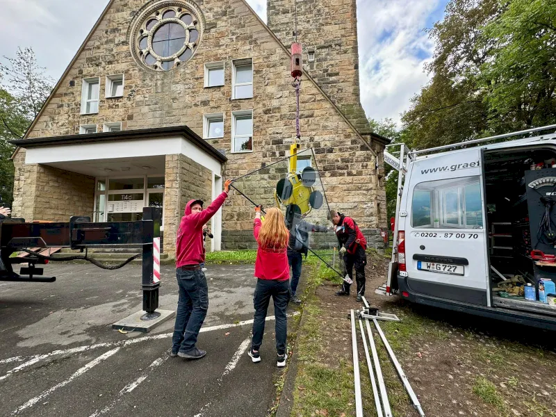 In Wuppertal führen drei Personen eine große Glasfensterscheibe, die von einem Kran vor ein steinernes Gebäude mit einem runden Fenster gehoben wird. Ein Lieferwagen mit Ausrüstung ist in der Nähe geparkt, und ein weiterer Arbeiter steht in der Nähe des Gebäudeeingangs.