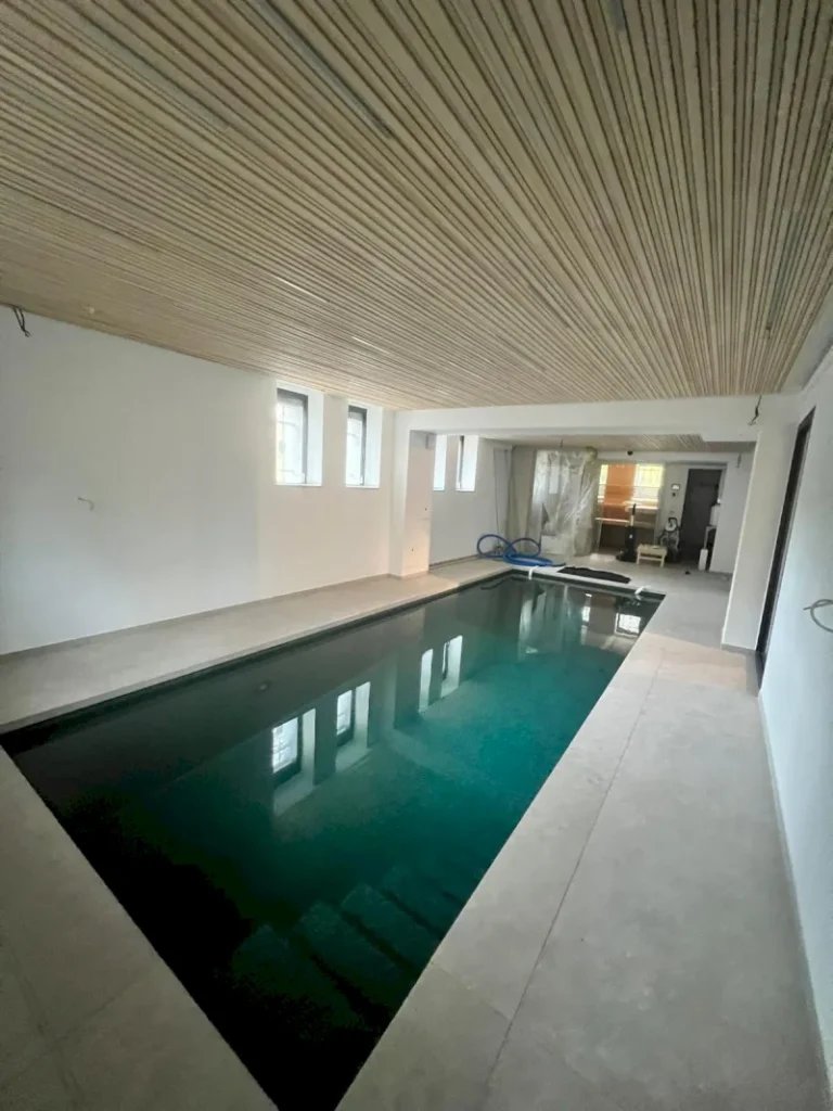 Ein Hallenbad mit klarem Wasser, beigem Fliesenboden und einer Holzlattendecke. Die weißen Wände und Glasfensterscheiben füllen den Wuppertaler Raum mit Licht, während an einem Ende die Beckenreinigungsgeräte stehen.