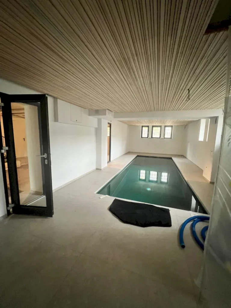 Ein modernes Hallenbad in einem weißen Raum mit einer Holzlattendecke, Glasfensterscheiben am anderen Ende und einer offenen Glastür. Poolreinigungsgeräte und eine schwarze Matte liegen auf dem Boden daneben und schaffen einen ruhigen Rückzugsort in Wuppertal.