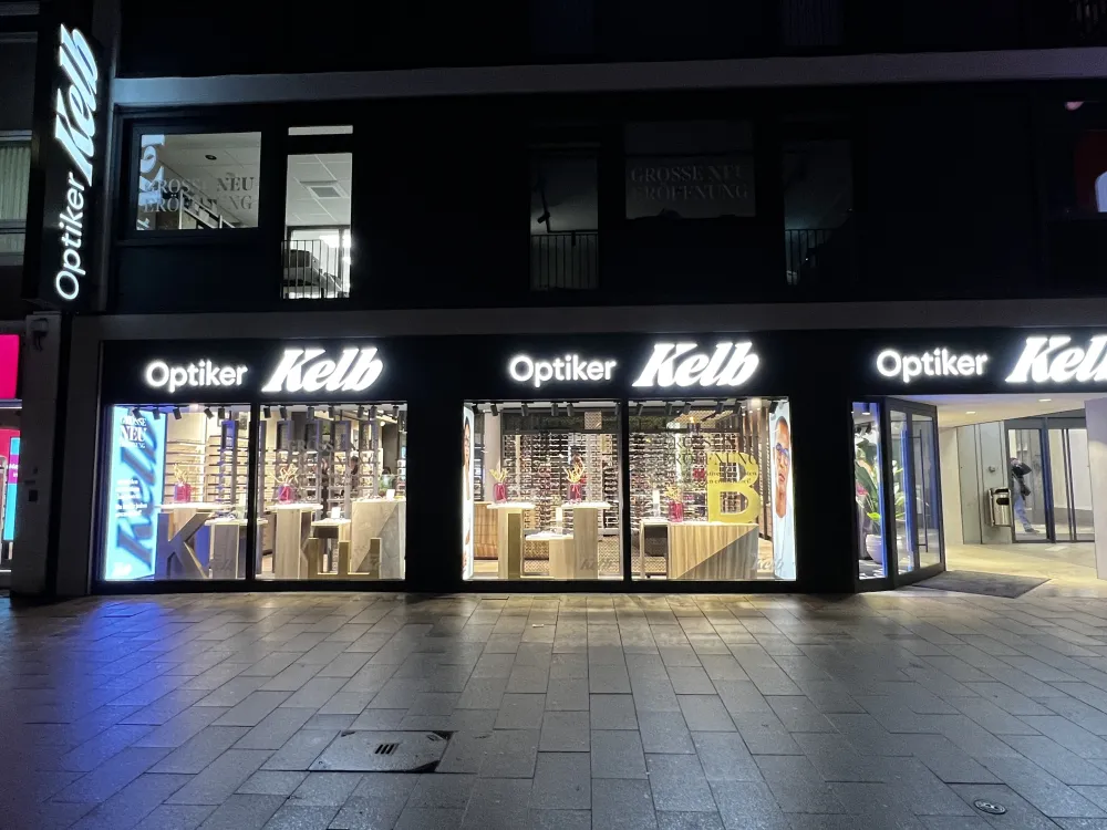 Ein hell erleuchtetes Optikergeschäft namens Optiker Kelb in Düsseldorf bei Nacht, mit großen Schaufensteranlagen für Brillen und Leuchtreklame über dem Eingang in einer ruhigen Straße.