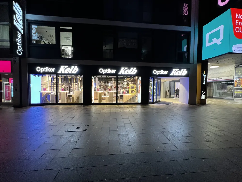 Ein hell erleuchtetes Brillengeschäft von Optiker Kelb in Düsseldorf bei Nacht, mit großen beleuchteten Schildern und gläsernen Schaufensteranlagen mit Brillenfassungen. Die steingepflasterte Straße davor ist leer und ruhig.