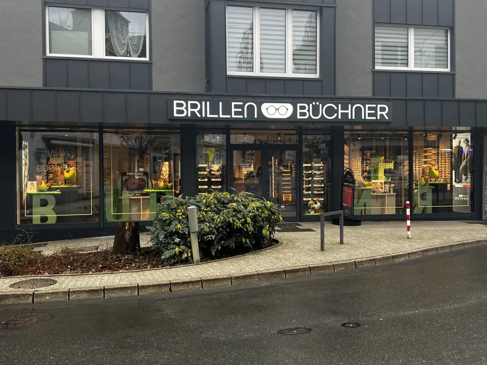 Ein Schaufenster von Brillen Büchner, einem Brillengeschäft in Düsseldorf, mit großen Schaufensteranlagen für Brillen und heller Beschilderung. Das graue Gebäude hat einen Gehweg, einige Sträucher und einen nassen Bürgersteig davor.