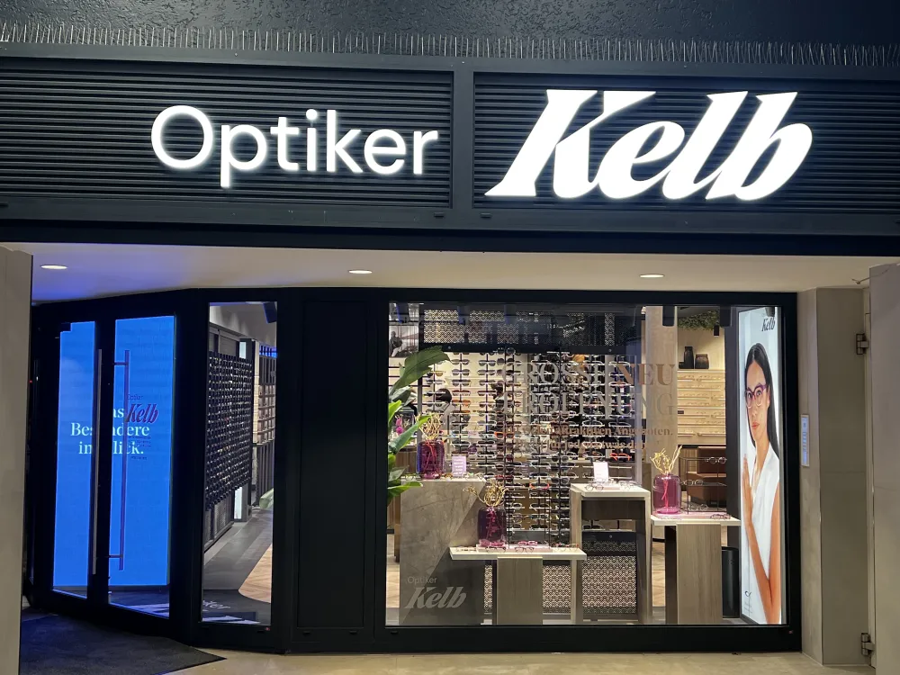 Ein Schaufenster in Düsseldorf bei Nacht zeigt das Schild Optiker Kelb. In den Schaufensteranlagen sind reihenweise Brillen und ein großes Plakat mit einer Brillenträgerin zu sehen. Die Eingangstür ist offen, im Inneren ist ein blauer Bildschirm zu sehen.