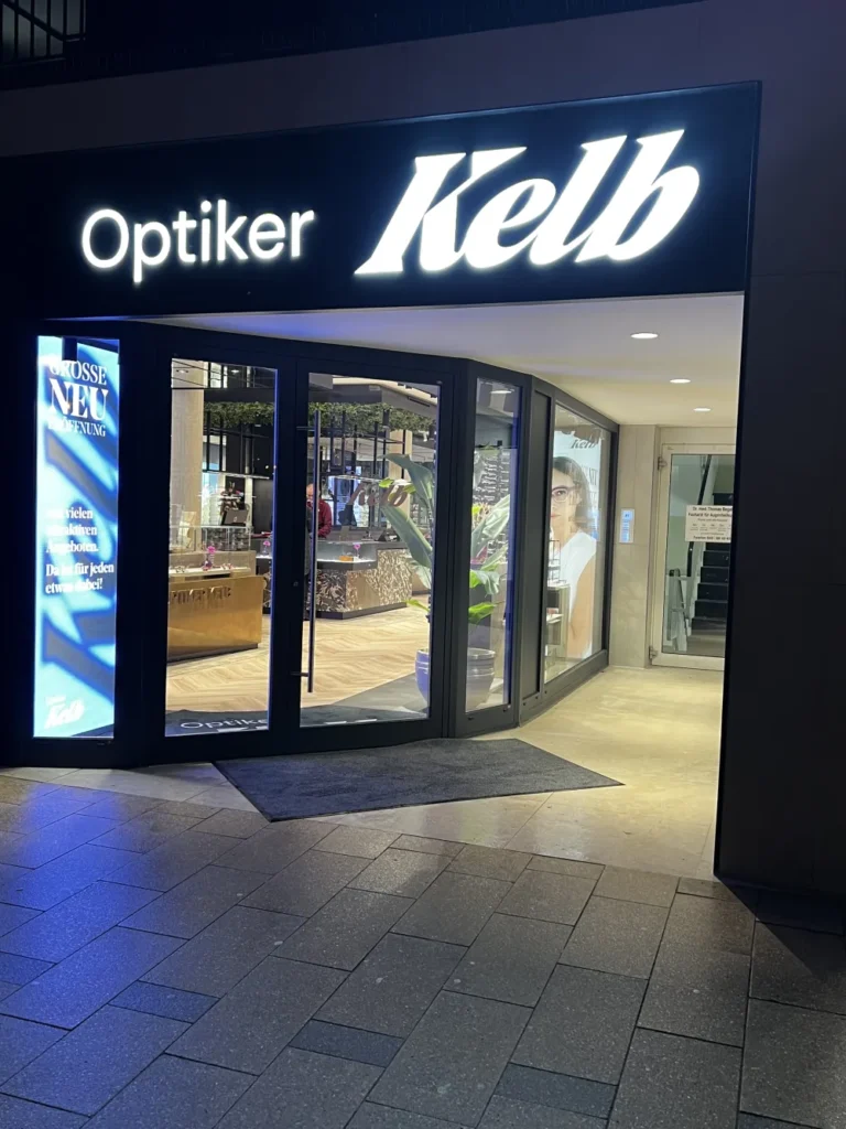 Die nachts hell erleuchtete Schaufensterfront mit dem Schriftzug Optiker Kelb über dem Eingang und die Glastüren mit der Schaufensterverglasung Düsseldorf zeigen ein modernes Interieur und große Plakate im Inneren.