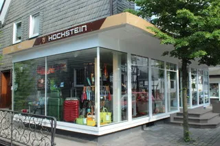 Ein modernes Eckgeschäft mit großen Glasfenstern bietet bunte Gepäckstücke, Taschen und Haushaltsartikel an. Auf dem Schild darüber steht "Hochstein" und zeigt die stilvollen Schaufensteranlagen Wuppertals. Ein kleiner Baum steht neben dem Eingang.