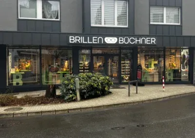 Ein Schaufenster mit großen Schaufensteranlagen in Wuppertal stellt Brillen aus. Über dem Eingang befindet sich ein Schild mit der Aufschrift BRILLEN BÜCHNER und einem Brillensymbol zwischen den Wörtern. Die nasse Straße davor deutet auf kürzlichen Regen hin.