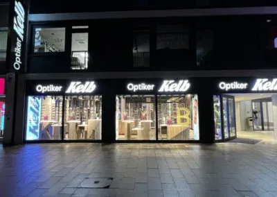 Ein gut beleuchtetes Schaufenster bei Nacht zeigt das Schild Optiker Kelb über großen Schaufensteranlagen, in denen Brillen und optische Auslagen zu sehen sind. Der Bürgersteig davor ist leer, was die ruhige Atmosphäre der Schaufensteranlagen Wuppertal widerspiegelt.