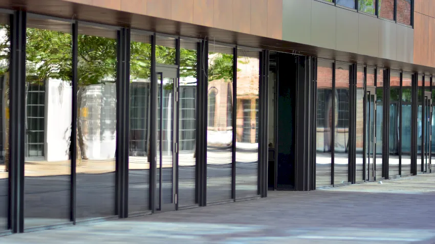 In einer Reihe hoher gläserner Schaufensteranlagen spiegeln sich Bäume und Gebäude auf einem modernen Geschäftshaus, wobei das Sonnenlicht Schatten auf den Gehweg davor wirft.
