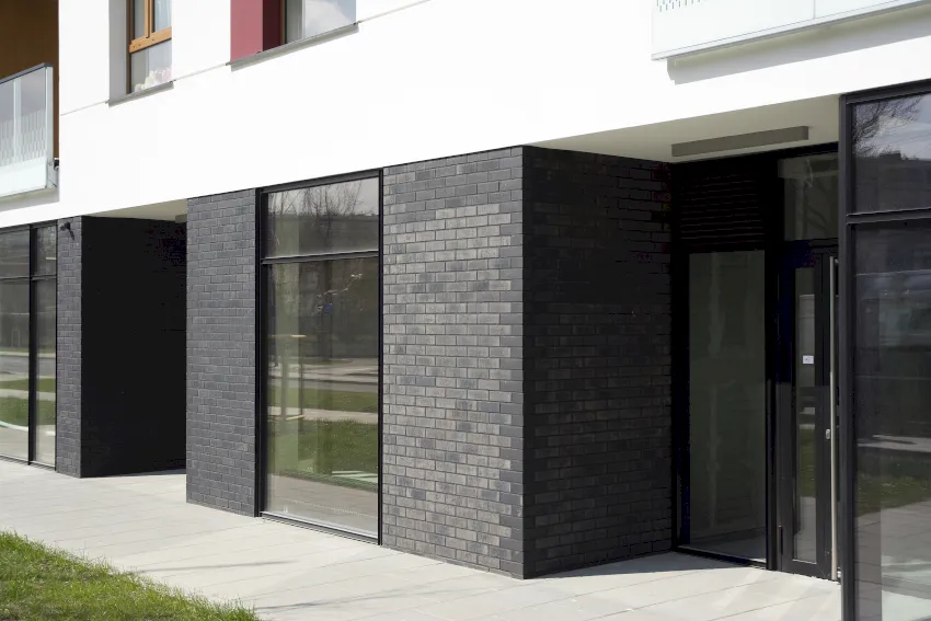Moderne Gebäudefassade mit großen Glasfenstern, perfekt für Schaufensteranlagen Wuppertal, schwarze Backsteinwände im Erdgeschoss, weißes Obergeschoss und Balkone. Ein gepflasterter Gehweg und ein grüner Grünstreifen vervollständigen das Bild.